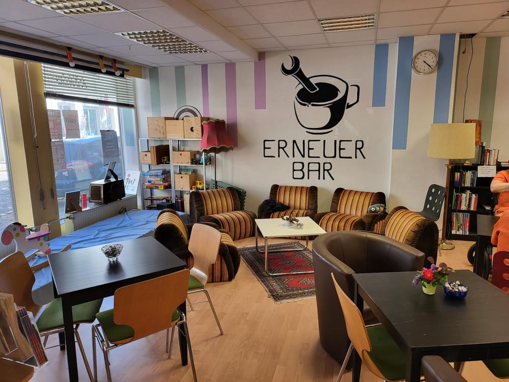 Bild der Erneuer-Bar. Zu sehen sind Tische mit Stühlen sowie eine Sitzgruppe aus 4 Sesseln mit einem Tisch. Im Hintergrund an der Wand ist eine Kaffeetasse mit einem Schraubenschlüssel
